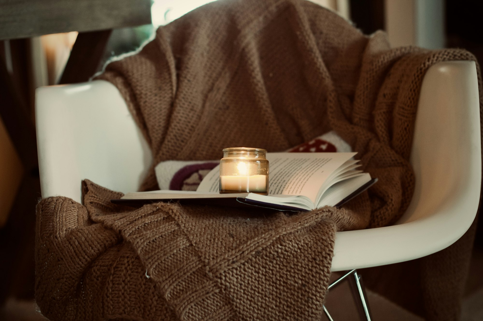An open book and small candle resting on a brown knit throw draped across a white chair.