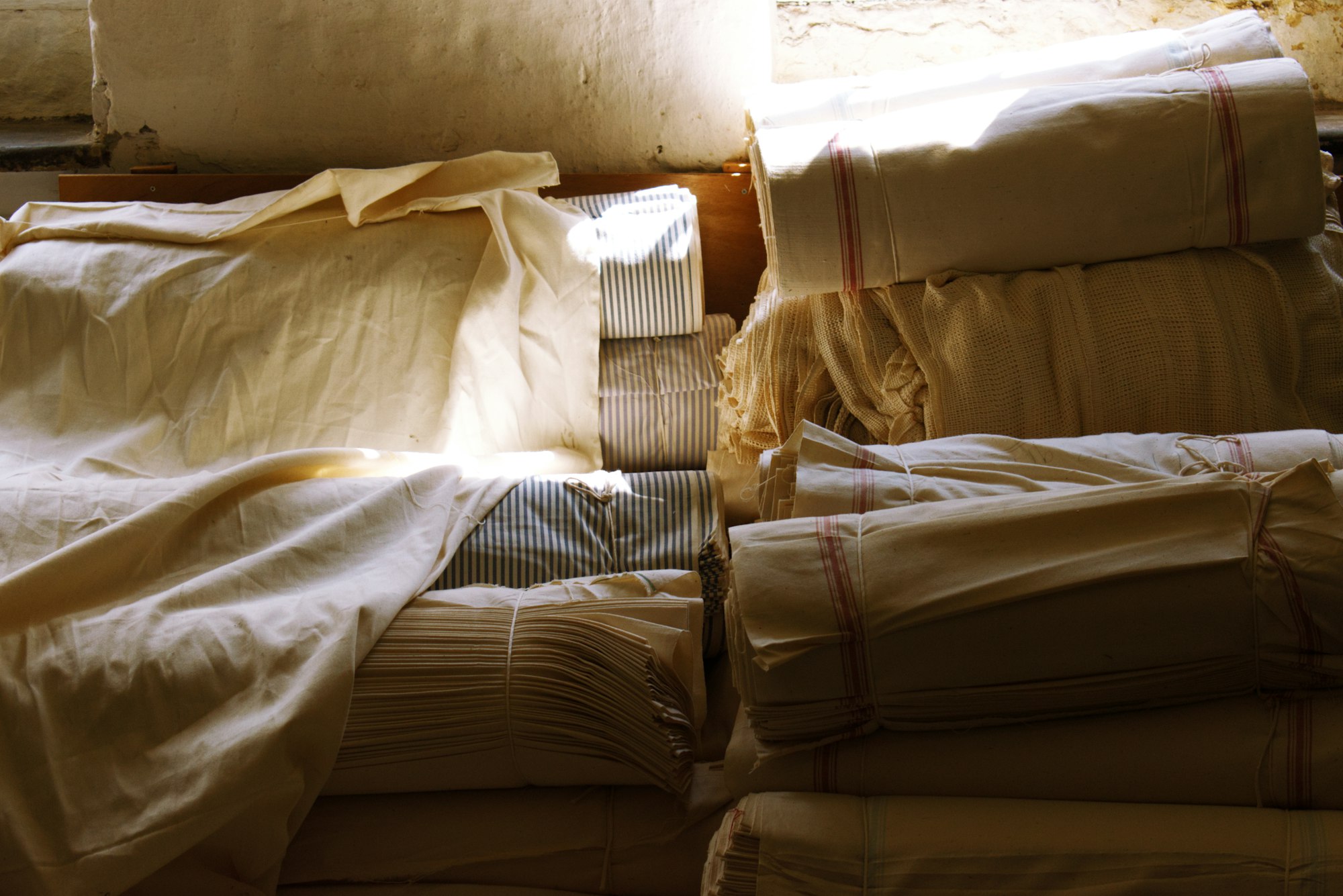 Folded cream linen sheets stacked on a wooden shelf, warm window light falling across them.